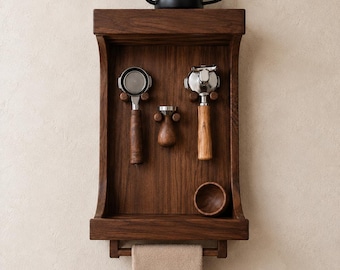 May include: A dark wood wall-mounted organiser featuring a top shelf holding a black gooseneck kettle. The rack displays two espresso portafilters, a wooden tamper, and a small bowl. A beige towel with the word ESPRESSO hangs from the bottom bar.