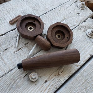 Pode incluir: Dois puxadores de madeira castanho-escuro com bordas octogonais e inserções quadradas de latão no centro, acompanhados por um cabo cilíndrico longo de madeira com uma extremidade de parafuso de metal. Uma pequena etiqueta de madeira diz NERO COFFEE.