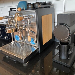 Puede incluir: Máquina de espresso de acero inoxidable con detalles de madera y medidores azules. Un molinillo de café negro con una taza negra está al lado de la máquina de espresso. Se ve una tostadora negra.