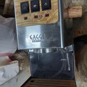 Puede incluir: Máquina de espresso Gaggia Classic de acero inoxidable con mango y panel de control de madera. El panel de control tiene tres botones negros con iconos y tres luces indicadoras rojas. La máquina tiene una bandeja de goteo y un depósito de agua negros.