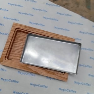 Könnte beinhalten: Ein Holzserviertablett mit einem rechteckigen Einsatz aus Edelstahl. Das Tablett besteht aus hellbraunem Holz mit einer geschnitzten Nut. Der Einsatz ist ein glänzender, rechteckiger Metallbehälter. Der Hintergrund hat ein sich wiederholendes Muster mit dem Text "NepoCoffee".