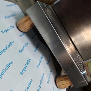 Op de afbeelding: Een roestvrijstalen koffietamper met houten handvatten ligt op papier met de blauwe tekst "NepoCoffee". De stamper is rechthoekig met een platte metalen basis.
