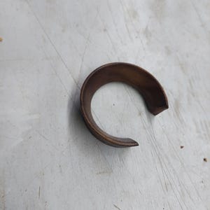 May include: A curved, bronze-colored metal piece, possibly a component or part of a larger mechanism. The object has a semi-circular shape with a visible gap at one end. The surface appears worn.