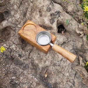 May include: A wooden espresso machine portafilter holder with a matching handle. The holder is made of light-colored wood and has a circular indentation to hold the portafilter. The portafilter is stainless steel.