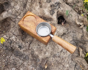 May include: A rectangular wooden block with a circular indentation and a stainless steel espresso portafilter basket featuring a matching wooden handle. The wood grain is visible throughout the block and the handle, showcasing natural light brown tones.