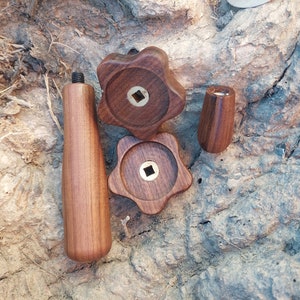 May include: A selection of wooden hardware components, including two star-shaped knobs with brass inserts, a cylindrical knob, and a threaded handle. The items are made of a rich, dark wood and are displayed on a textured, light brown surface.