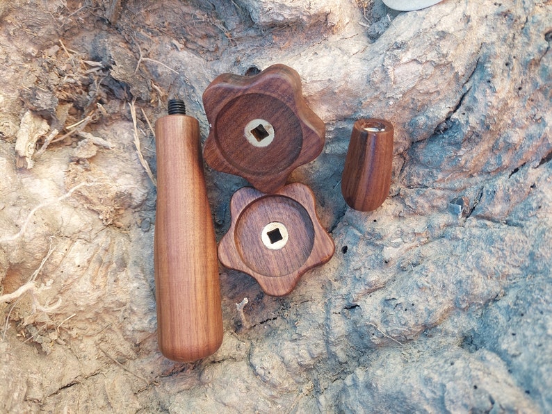 May include: A selection of wooden hardware components, including two star-shaped knobs with brass inserts, a cylindrical knob, and a threaded handle. The items are made of a rich, dark wood and are displayed on a textured, light brown surface.