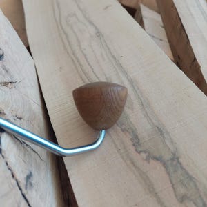 May include: A wooden handle with a rounded top and a curved metal hook. The handle is a light brown colour and the metal hook is silver. The handle rests on a light brown wooden surface.