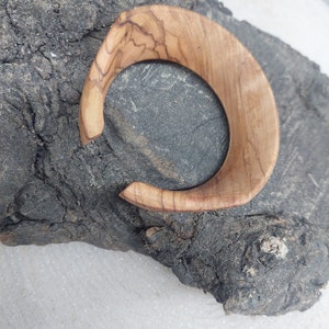 May include: A wooden bangle with a natural wood grain pattern. The bangle is light brown in color and has a smooth, polished finish.