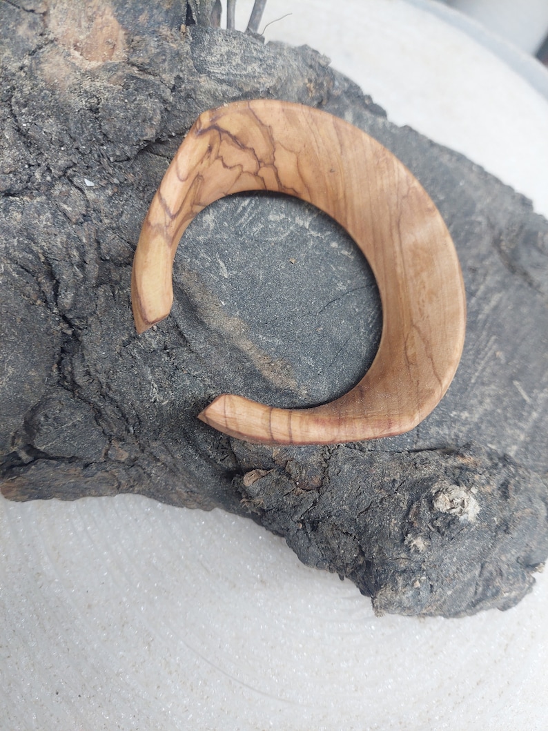 May include: A wooden bangle with a natural wood grain pattern. The bangle is light brown in color and has a smooth, polished finish.