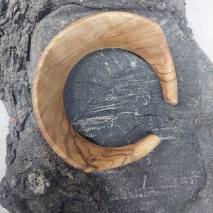 May include: A wooden bangle bracelet with a natural wood grain finish. The bracelet is a light brown color and has a smooth, polished surface.