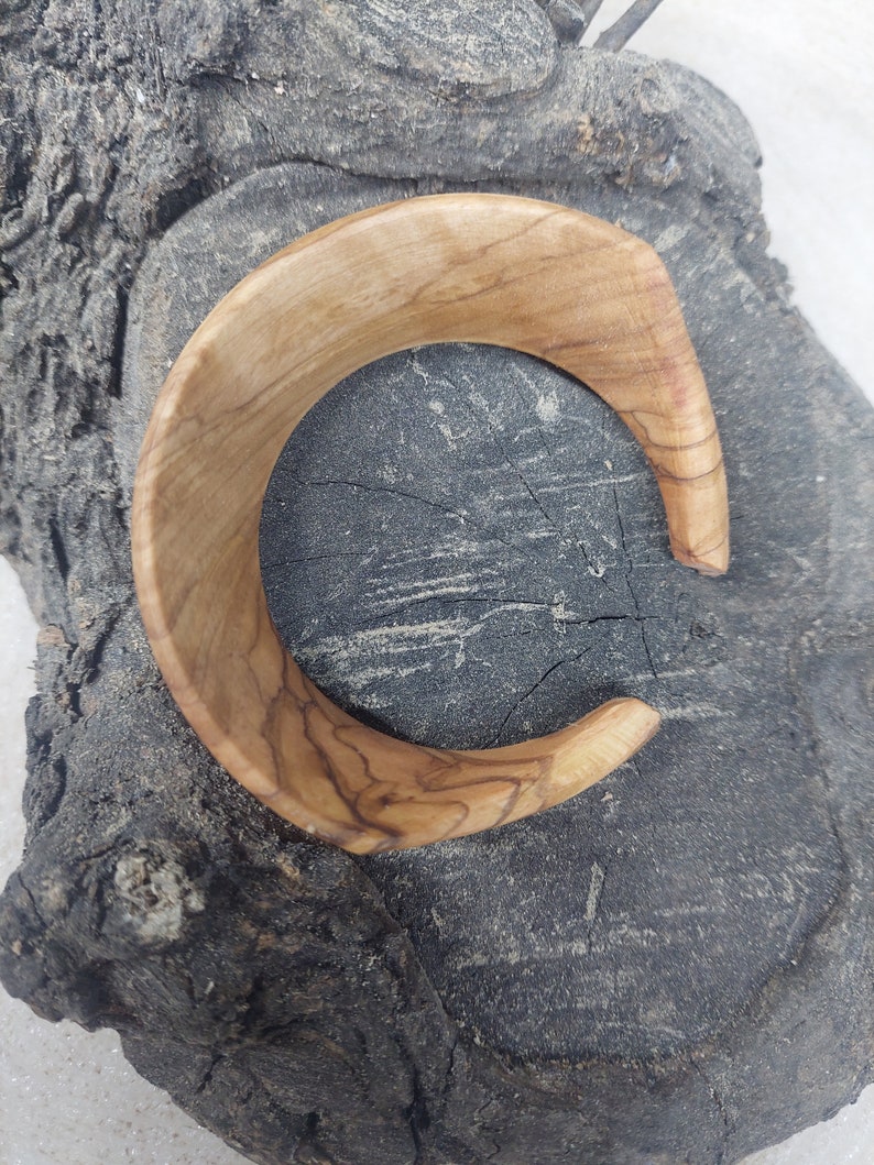 May include: A wooden bangle bracelet with a natural wood grain finish. The bracelet is a light brown color and has a smooth, polished surface.