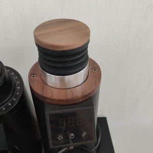 Könnte beinhalten: Eine schwarze elektrische Kaffeemühle mit einer Holzplatte und einem digitalen Display, das "8.8.8" anzeigt. Die Mühle hat einen silbernen Ring um die Oberseite und einen schwarzen Becherhalter.