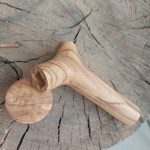 Puede incluir: Un tirachinas de madera de olivo con un acabado de veta de madera natural. El tirachinas tiene un mango en forma de Y y una base redonda. El tirachinas está apoyado sobre una superficie de madera.