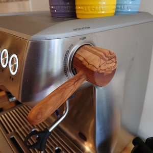 Könnte beinhalten: Eine Espressomaschine aus Edelstahl mit einem Dampfstabgriff aus Holz. Der Griff ist aus Olivenholz gefertigt und hat ein natürliches Maserungsmuster. Die Maschine hat ein Zifferblatt mit der Aufschrift "STEAM" und "OPEN".