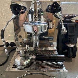 May include: A polished stainless steel espresso machine with a black handle and two pressure gauges. The machine features a portafilter and two steam wands. A coffee grinder is in the background.