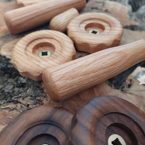 Puede incluir: Una colección de pomos de madera de diferentes formas y tamaños. Los pomos están hechos de madera clara y oscura y tienen un agujero cuadrado en el centro. Los pomos están dispuestos sobre una superficie de madera.