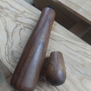 Puede incluir: Un tapón de botella de madera con una parte superior redondeada y una base cilíndrica. El tapón está hecho de madera marrón oscura con un acabado liso.