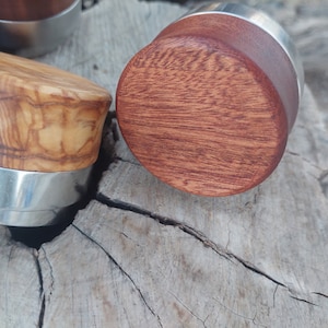 Könnte beinhalten: Zwei Flaschenverschlüsse aus Holz mit Metallsockeln. Ein Verschluss ist aus hellbraunem Holz mit einem wirbelnden Maserungsmuster. Der andere Verschluss ist aus dunkelrotem Holz mit einem geraden Maserungsmuster.