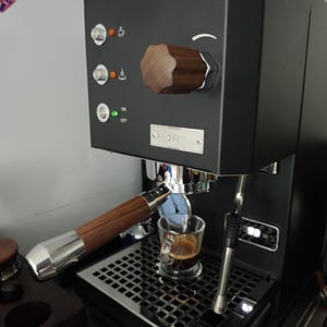 May include: Black espresso machine with a wooden handle and knob. A small glass cup is filled with espresso. The machine has the brand name "Profitec" on the front.