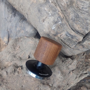May include: A coffee tamper with a wooden handle and a black base with a silver rim. The word "normcore" is printed on the base. The tamper is resting on a piece of weathered wood.