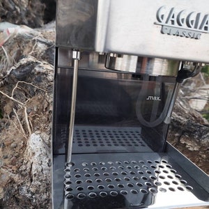 Puede incluir: Una máquina de espresso Gaggia Classic de acero inoxidable con base de madera. La máquina tiene una bandeja de goteo con un inserto de metal perforado y un tanque de agua con una línea de llenado máxima.