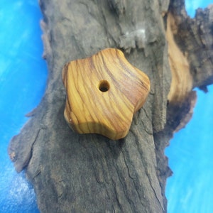May include: A star-shaped wooden bead with a hole in the centre. The bead is made of light brown wood with darker brown grain patterns. The bead is resting on a piece of dark grey wood.