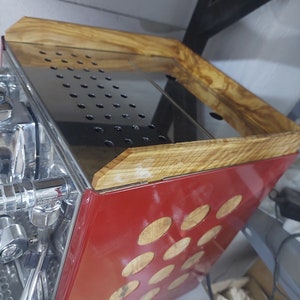 Puede incluir: Una máquina de espresso roja con una tapa de madera y una bandeja de goteo de acero inoxidable. La máquina tiene un acabado cromado y una serie de agujeros redondos en el lateral.