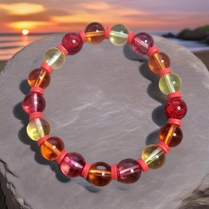 May include: A colourful beaded bracelet with translucent beads in red, orange, and yellow. The beads are separated by small, coral-coloured spacers. The bracelet is displayed on a grey, textured surface with a sunset in the background.