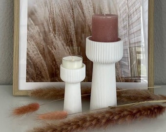 May include: Two white ceramic candle holders with a ribbed design. The larger holder has a pink candle inside. The smaller holder has a white candle inside. The candles are in front of a framed print with the word "DREAM" on it. The print has a background of brown and white dried grass.