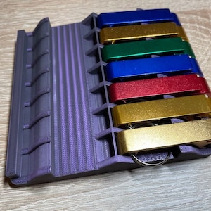 Puede incluir: Un soporte de color morado impreso en 3D con siete ranuras para abridores de botellas. Las ranuras están llenas de abridores de botellas de varios colores, incluyendo dorado, rojo, azul y verde.