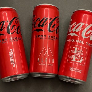 Könnte beinhalten: Drei rote Aluminium-Coca-Cola-Dosen mit unterschiedlichen Etiketten. Die linke Dose trägt den Schriftzug "Zero Sugar, Special Edition for Greta". Die mittlere Dose trägt den Schriftzug "Zero Sugar, Alpin, Kulinarik Hotel". Die rechte Dose trägt den Schriftzug "Original Taste, Lenny, Delicious & Refreshing".