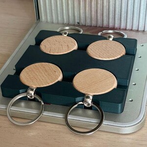 May include: Four wooden keychains with metal rings are displayed in a black plastic mold. The keychains are round and have a natural wood finish.