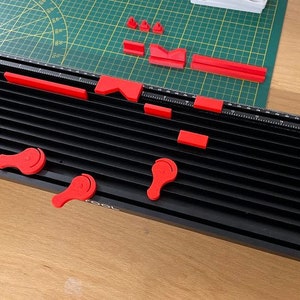 May include: A black and red tool with multiple components. The main part is a black, grooved base with a ruler along the edge. Red pieces, including handles and rectangular blocks, are scattered around the base. A green cutting mat is in the background.