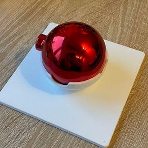 Könnte beinhalten: Eine glänzende rote Weihnachtskugel mit reflektierender Oberfläche, die auf einer weißen quadratischen Basis liegt. Der Schmuck hat eine kleine, zylindrische Oberseite zum Aufhängen. Das Design ist schlicht und festlich.