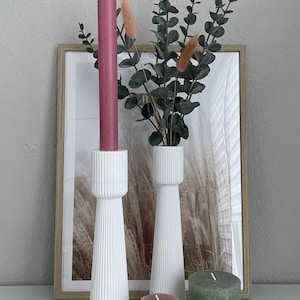 Könnte beinhalten: Drei weiße Kerzenhalter mit geripptem Design, zwei mit rosa und grünen Kerzen und einer mit einer rosa Kerze. Die Kerzenhalter stehen vor einem gerahmten Bild mit einem braunen und weißen abstrakten Design. Ein Strauß grüner Eukalyptuszweige befindet sich in der Mitte des Bildes.