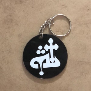 Pode incluir: Chaveiro redondo preto com corrente e argola prateadas. O chaveiro apresenta caligrafia árabe branca e um desenho de cruz. O fundo é uma superfície castanha clara.