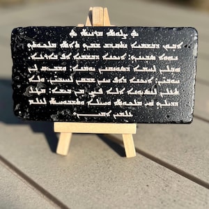 Può includere: Una placca rettangolare nera con testo bianco, esposta su un piccolo cavalletto di legno. Il testo è una preghiera in una scrittura antica. La placca ha una superficie strutturata e si trova su uno sfondo esterno sfocato.