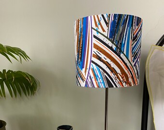 May include: A black and silver table lamp with a white lampshade featuring a colourful abstract design in shades of blue, orange, and black. The lamp is on a glass table top with a blue and white crocheted basket.