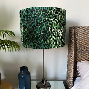 Op de afbeelding: Een groene en zwarte lampenkap met luipaardprint met een zwarte metalen lampvoet. De lamp staat op een glazen tafel met een rieten mand en een blauwe vaas.