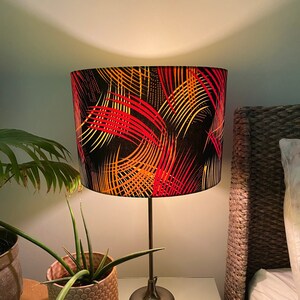May include: A table lamp with a black lampshade featuring a vibrant pattern of red, orange, and yellow curved lines. The lamp sits on a mirrored surface, next to potted plants and a bed.
