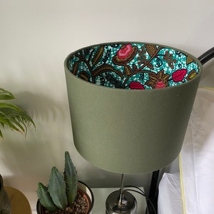 Op de afbeelding: Een groene lampenkap met een bloemenpatroon aan de binnenkant. De lampenkap staat op een tafel naast een cactus in een pot.
