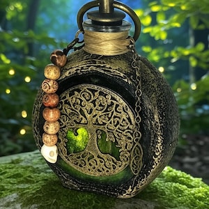 Pode incluir: Uma garrafa de vidro redonda e decorativa com um design de árvore da vida em metal cor de bronze. A garrafa tem um exterior escuro e texturizado com detalhes dourados, um anel de bronze e uma corrente de contas. O interior brilha em verde.