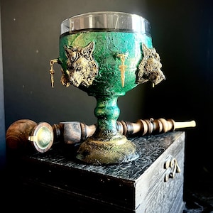 Pode incluir: Um cálice decorativo com uma taça de vidro transparente e uma base pintada de verde e dourado. O cálice é adornado com detalhes de cabeça de lobo dourados e pequenos amuletos pendurados. Um cachimbo de madeira e uma caixa de madeira também estão na imagem.