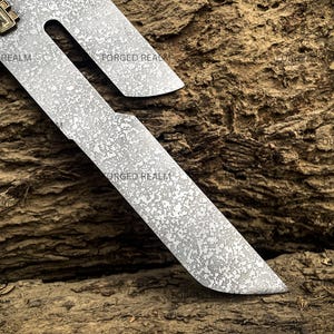 May include: A metal blade featuring a distinct mottled, textured surface pattern and a cutout section near the top. A small, rectangular brass-colored component is attached to the upper edge. The text FORGED REALM is visible on the blade.