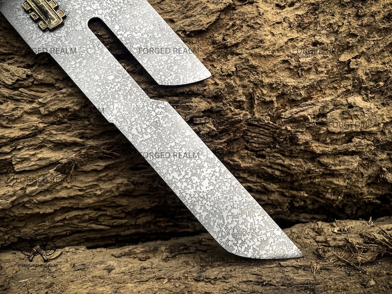May include: A metal blade featuring a distinct mottled, textured surface pattern and a cutout section near the top. A small, rectangular brass-colored component is attached to the upper edge. The text FORGED REALM is visible on the blade.