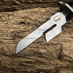 May include: A fixed-blade knife with a patterned steel blade featuring a cutout design and a small gold-colored rectangular inlay. The handle is dark brown with a metallic guard. The blade surface has a mottled texture and the text FORGED REALM.