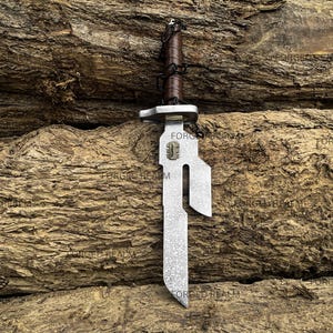 May include: A metal dagger featuring a textured silver blade with a distinct cutout notch and a small rectangular emblem. The handle is wrapped in dark brown leather with a black cord lacing detail and a silver crossguard separating the blade.