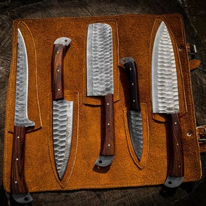 May include: A set of five kitchen knives with dark wood handles and silver-toned, hammered metal blades. The knives are arranged in a tan leather roll-up case with individual slots for each blade, featuring metal rivets and a buckle closure.