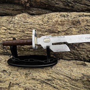 May include: A metal knife with a brown leather-wrapped handle, a brass-colored rectangular accent on the blade, and a cutout section near the hilt. The knife rests on a black oval stand with a decorative chain, featuring the text FORGED REALM.